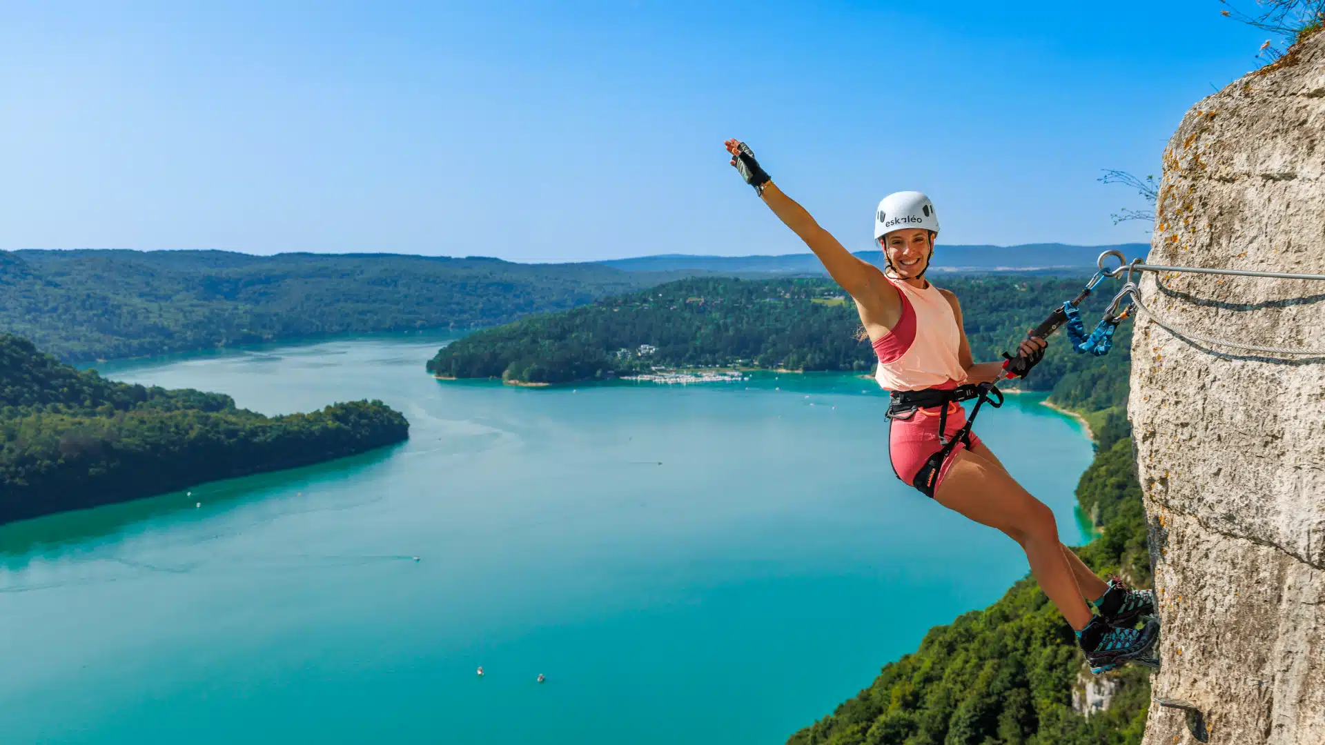 Une personne escaladant une via ferrata dans le Jura avec vue panoramique sur le lac de Vouglans